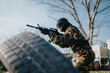 © qunica.com - A soldier in camouflage armor crouches and aims a rifle behind a tire obstacle. The scene shows a military training moment in an outdoor setting.