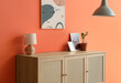 © Pixel-Shot - Chest of drawers with lamp, magazine and houseplant near orange wall in living room, closeup