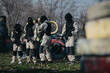 © qunica.com - A group of players in full paintball gear stands ready with markers. They gather on an obstacle-filled outdoor field for a game.