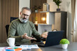 © Dejan - Mature man feeling frustrated and stressed, gesturing confusedly while looking at a laptop computer in a contemporary home office