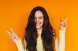 © zvkate - Portrait of playful young woman showing peace sign gesture on orange background, happy girl with long hair making V-sign and smiling, positive vibes and youthful lifestyle concept, closed eyes