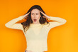 © zvkate - Frustrated young woman holding head in hands with annoyed expression on orange background, angry girl shouting in disbelief or failure, stress and negative emotions concept, studio portrait