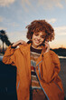 © SHOTPRIME STUDIO - Smiling woman with curly hair wearing rainbow sweater and orange jacket talks on smartphone outdoors during sunset with blurred background and positive lifestyle mood.