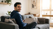 © Milan - Young man working on laptop on sofa at home. Smiling male freelancer looking back in bright modern living room. Remote work and digital nomad lifestyle concept. Copy space for text