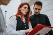 © qunica.com - Red-haired woman in a vest and glasses presents a document to colleagues. They review the red folder outside a modern building.