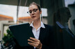 © BullRun - Caucasian businesswoman in glasses reading tablet screen attentively, standing near glass wall outdoors, showing focus, remote task processing and digital workspace engagement.