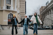 © qunica.com - Three girls walk together down a city street in winter, smiling in stylish coats. They hold hands and stroll past modern buildings on a cloudy day.