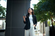 © Parichat - Young Asian businesswoman using smartphone looking sideways outdoors