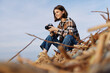 © SHOTPRIME STUDIO - Woman photographer crouches on driftwood by the shore, holding a camera and scanning the scene for the perfect shot as waves roll under a blue sky above the calm coastline.
