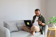 © ArvStd - An adult man sits on a sofa happily using a bank credit card and laptop while transferring money, making cashless online purchases, and shopping online for home delivery.