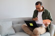 © ArvStd - Adult man sitting on sofa at home holding Indonesian banknotes, staring and pointing thoughtfully at laptop screen while deciding on expenses, considering purchases, investments or financial planning.