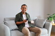 © ArvStd - Handsome Asian man with a cup of coffee and holding a cellphone, relaxing on the sofa at home, smiling at the camera, enjoying his free time at home.