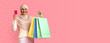 © Prostock-studio - A woman wearing a headscarf stands against a pastel background. She holds several shopping bags in one hand and a card in the other. She has a big smile on her face.
