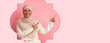 © Prostock-studio - A young woman wearing a light hijab points with both hands to an empty area. The background is pink and has a decorative shape. This scene is cheerful and invites creativity.