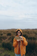 © SHOTPRIME STUDIO - Young woman lifestyle outdoors wearing orange jacket and glasses takes photo with smartphone in natural field under cloudy sky. Concept of modern technology and nature connection.