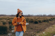 © SHOTPRIME STUDIO - Young woman lifestyle walking outdoors in autumn field wearing orange jacket and knit hat, holding smartphone and smiling. Concept of casual fashion, nature, and technology balance.