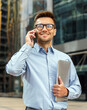 © muse studio - Businessman smiles while holding a laptop and talking on a cell phone in a modern city during the day