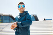 © muse studio - Active man in blue sportswear smiles, checking his smartwatch on an urban staircase during a bright sunny day