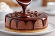© whitestorm - Pouring warm chocolate sauce over a decadent chocolate mousse cake, topped with chocolate chunks, on a white plate, with blurred background showcasing a culinary masterpiece.