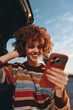 © SHOTPRIME STUDIO - Smiling woman with curly hair wearing a rainbow striped sweater looks at smartphone near car outdoors. Woman enjoys lifestyle moment with natural light and clear sky background.