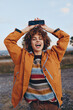 © SHOTPRIME STUDIO - Happy woman with curly hair wearing rainbow sweater and orange jacket smiles holding smartphone above head outdoors in casual lifestyle setting during daytime.