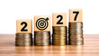 © Urmi - 'Wooden cubes with 2027 numbers on coin stacks illustrate saving money, investment growth, income goals, and business strategies for economic success.'
