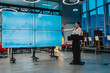 © qunica.com - A presenter speaks at a podium beside a multi-screen financial dashboard. The modern office features a data-driven briefing with charts and graphs.
