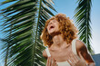 © SHOTPRIME STUDIO - Laughing woman with curly hair enjoying sunny tropical outdoors near palm leaves, dressed in white tank top under clear blue sky with bright sunlight.