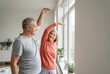 © Tima - Senior man and senior woman exercising by window at home. Elderly couple doing stretching workout for healthy lifestyle. Active retirement, physical activity and wellness concept.