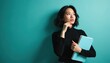 © Raisa - Asian woman in black turtleneck holds blue notebook, looks up thoughtfully. She has chin resting on hand, considering idea. Indoor studio backdrop.