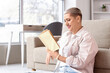 © Pixel-Shot - Happy mature blonde woman with short hair reading book on floor at home