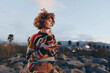 © SHOTPRIME STUDIO - Young woman with curly hair wearing a colorful rainbow sweater stands outdoors on rocky terrain, holding a smartphone, smiling thoughtfully, lifestyle concept with natural background and soft light.