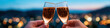 © Sweettymojidesign - Two hands holding sparkling champagne glasses toasting with blurred city lights in background creating festive and joyful atmosphere at dusk