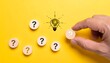 © EPDICAY - Hand holding a wooden token with a question mark near a glowing light bulb icon on a yellow background, symbolizing problem solving, ideas, and decision making.
