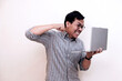 © Nurma - Southeast Asian man wearing flannel shirt standing and holding laptop with angry expression, on white isolated background.  Ideal for content about success, tech joy, or breakthrough moments