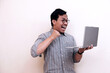 © Nurma - Southeast Asian man wearing flannel shirt standing and holding laptop with angry expression, on white isolated background.  Ideal for content about success, tech joy, or breakthrough moments