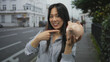 © Krakenimages.com - Young woman outdoors holding a piggy bank, smiling with a thumbs-up gesture in an urban street setting, emphasizing saving money and positive financial habits.