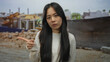 © Krakenimages.com - Young chinese woman standing at a construction site outdoors with a serious expression, surrounded by building equipment and dirt, wearing a casual white shirt.