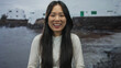 © Krakenimages.com - Young chinese woman smiling happily by the seaside with white buildings in the background on a tranquil beach day outdoors.