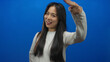 © Krakenimages.com - Young chinese woman saluting with fingers on head stands against isolated blue background wall showcasing playful confident expression.