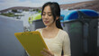 © Krakenimages.com - Woman holding clipboard outdoors near recycling containers with focus on sustainable waste management in a sunny urban setting.