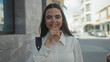 © Krakenimages.com - Woman outdoors smiling on an urban street, wearing white shirt, illustrating quiet with finger gesture, bright daytime setting.