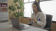© Krakenimages.com - Hispanic woman doctor with stethoscope conducting a video consultation in an office surrounded by medical equipment and greenery, showcasing a modern clinical workplace.