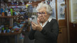 © Krakenimages.com - Elderly woman wearing glasses holding smartphone, finger near mouth in a cluttered retail shop building; concentration.