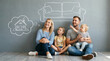 © Mr - Family of four sits on a wooden floor against a gray wall, dreaming of a new house and a comfortable sofa drawn in chalk above them.