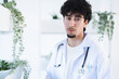 © Studio Marmellata - A young male doctor wearing a white coat and stethoscope stands in a bright, modern clinic, surrounded by plants.