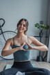 © Daniel - Young woman sitting on a yoga mat at home, smiling at the camera and forming a heart shape with her hands in front of her chest, radiating positivity, self-love, and inner happiness