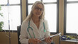 © Krakenimages.com - Young woman doctor with blonde hair wearing a stethoscope and glasses writes notes on a clipboard in a modern clinic room filled with natural light and medical equipment.