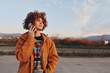 © SHOTPRIME STUDIO - A smiling woman in a rainbow sweater and orange jacket talks on a smartphone outdoors during golden hour with mountains in the background. The woman enjoys a casual lifestyle moment.