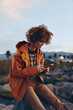 © SHOTPRIME STUDIO - A woman in an orange jacket and rainbow sweater smiles while using a smartphone outdoors during sunset. The woman enjoys a casual lifestyle in a natural rocky environment with mountains behind.